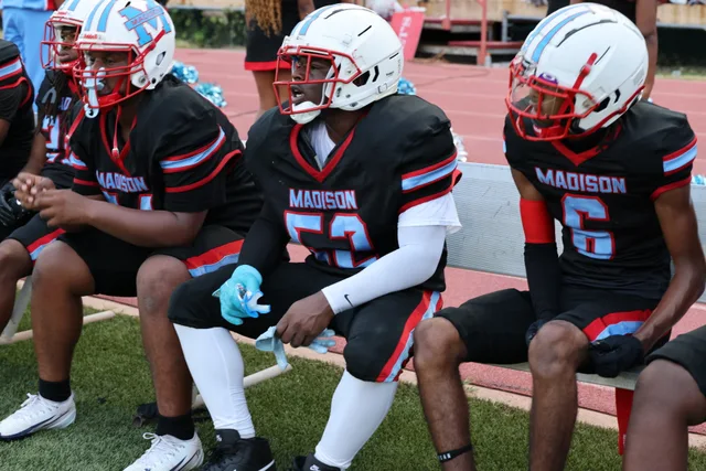 Madison players seated together on the sideline bench, wearing white helmets and black uniforms.