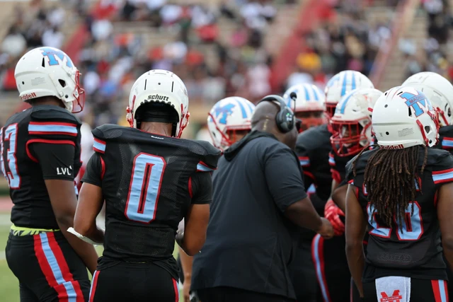 A coach speaks to a huddle of Madison players in black uniforms and white helmets.